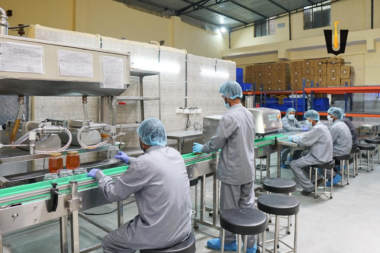 Workers filling glass honey jars on a green conveyor with a stainless multi-nozzle filling machine