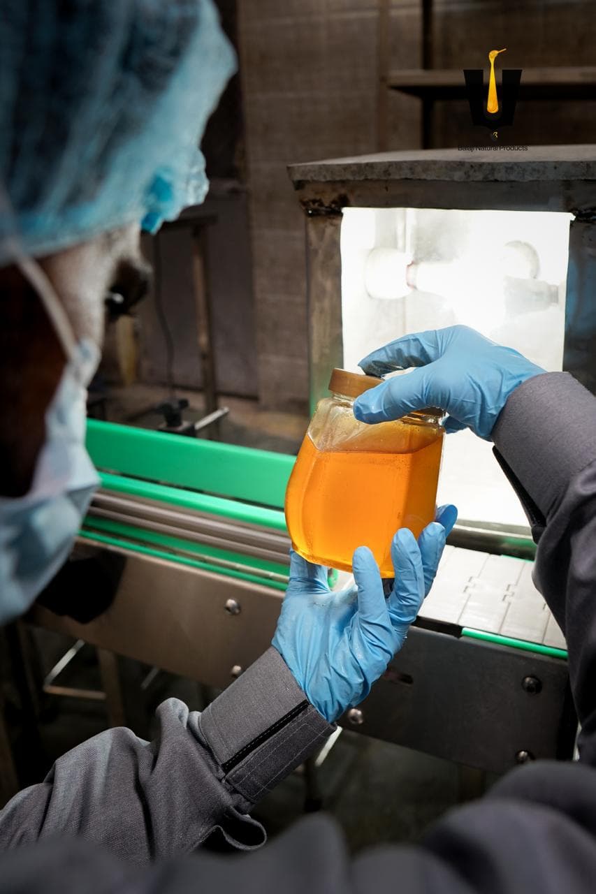 Worker in PPE inspecting a hexagonal jar of amber honey against a light box on the packing line
